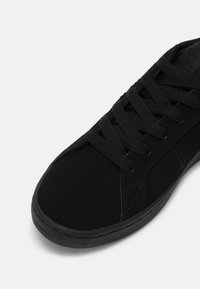 Zapatilla negra con una textura suave y mate, puntera redonda, cordones planos y un sutil detalle de costura en la parte superior y la suela.