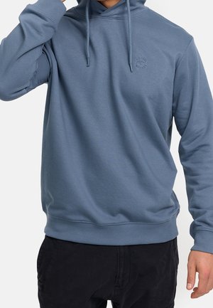 Homme portant un sweat à capuche bleu avec cordons et un pantalon noir, main près du visage, torse visible sur fond blanc.