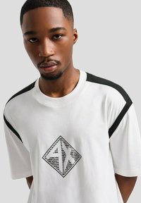 T-shirt bianco con dettagli neri sulle spalle, caratterizzata da un logo a forma di diamante con testo ripetuto in nero sul fronte. Tessuto morbido.