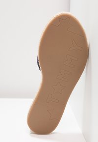Semelle en caoutchouc avec étoile inversée et "TOMMY" estampé. Accents en corde de jute naturelle le long du bord. Présente une surface beige texturée.