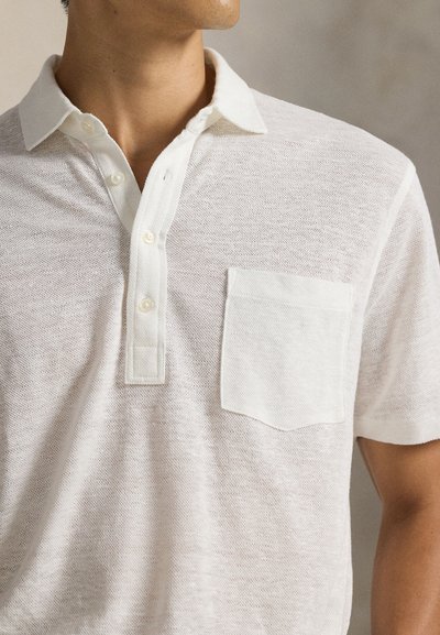 Κοντινό πλάνο ενός άνδρα με λευκό, υφασμάτινο κοντομάνικο πόλο πουκάμισο με μπροστινή τσέπη και τρία κουμπιά, να κοιτάζει προς τα πλάγια.