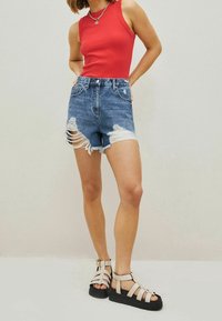 Red sleeveless top, high-waisted distressed denim shorts with frayed hem, and black platform sandals with beige straps. Plain background.
