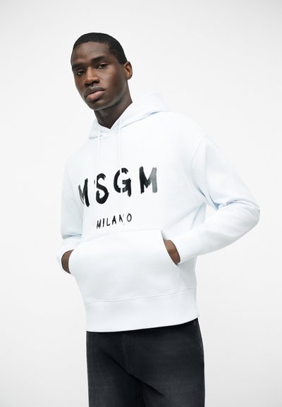 Νέος άνδρας που φοράει λευκό φούτερ MSGM Milano με τα χέρια στην μπροστινή τσέπη, ποζάρει μπροστά σε απλή λευκή φόντο.