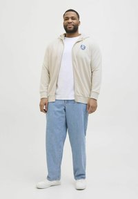 Svijetlosmeđi zip-up dukserica s logom, uparena s bijelom majicom i širokim plavim trapericama. Footwear čine bijele teniske. Jednostavan, ležeran outfit.