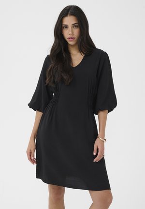 Woman with long dark hair wearing a black knee-length dress with puffed three-quarter sleeves against a plain background.