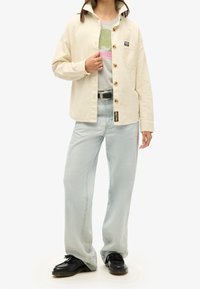 Beige kordfløjl skjortejakke med træknapper, båret over en grå grafisk t-shirt, parret med lyseblå lige ben jeans og sorte sko.