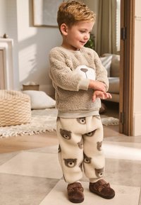 Fleece pajama set in beige with bear face design on top and bear patterns on pants. Soft texture and matching brown slippers.