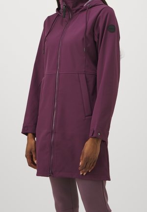 Femme portant une longue veste à capuche violette avec fermeture éclair à l'avant et poches latérales, associée à un pantalon violet assorti.