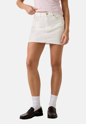 Donna che indossa una minigonna di denim bianca, calzini bianchi e mocassini neri, in piedi contro uno sfondo chiaro uniforme.