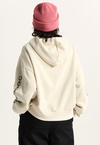 Romige hoodie met capuchon en een geribde zoom, voorzien van een kettinggrafiek op de linkermouw. Gecombineerd met zwarte broek en een roze gebreide muts.