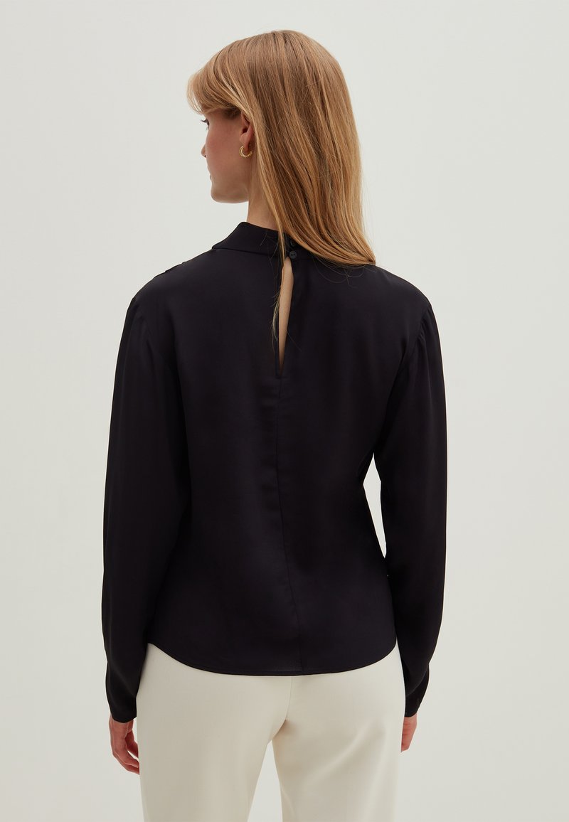 Zwarte blouse met lange mouwen, een hoge kraag en een keyhole-ontwerp aan de achterkant. Gladde textuur, iets getailleerde vorm en een discrete knoopsluiting bij de nek.