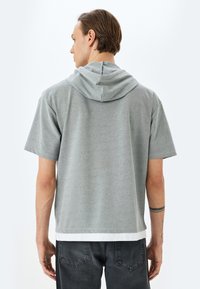 Sudadera gris de manga corta con cordón ajustable, tejido suave, corte relajado y detalle de dobladillo en capas sobre pantalones oscuros.