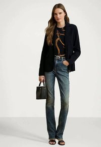 Žena v čiernom saku, vzorovanej čiernej blúzke, modrých džínsach, čiernych sandáloch, ktorá drží malú čiernu kabelku na jednotnom pozadí.