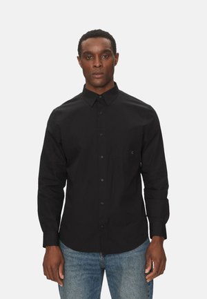 Camisa negra de manga larga abotonada con cuello, un bolsillo frontal y dobladillo recto, combinada con jeans azules.