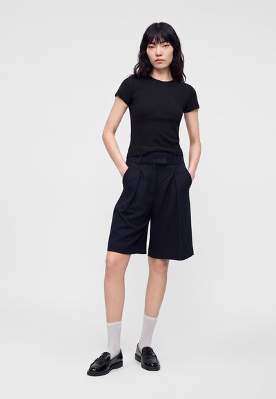 Femme aux longs cheveux noirs portant un haut noir côtelé à manches courtes, un short large noir arrivant aux genoux, des chaussettes blanches et des mocassins noirs, debout, les mains dans les poches.