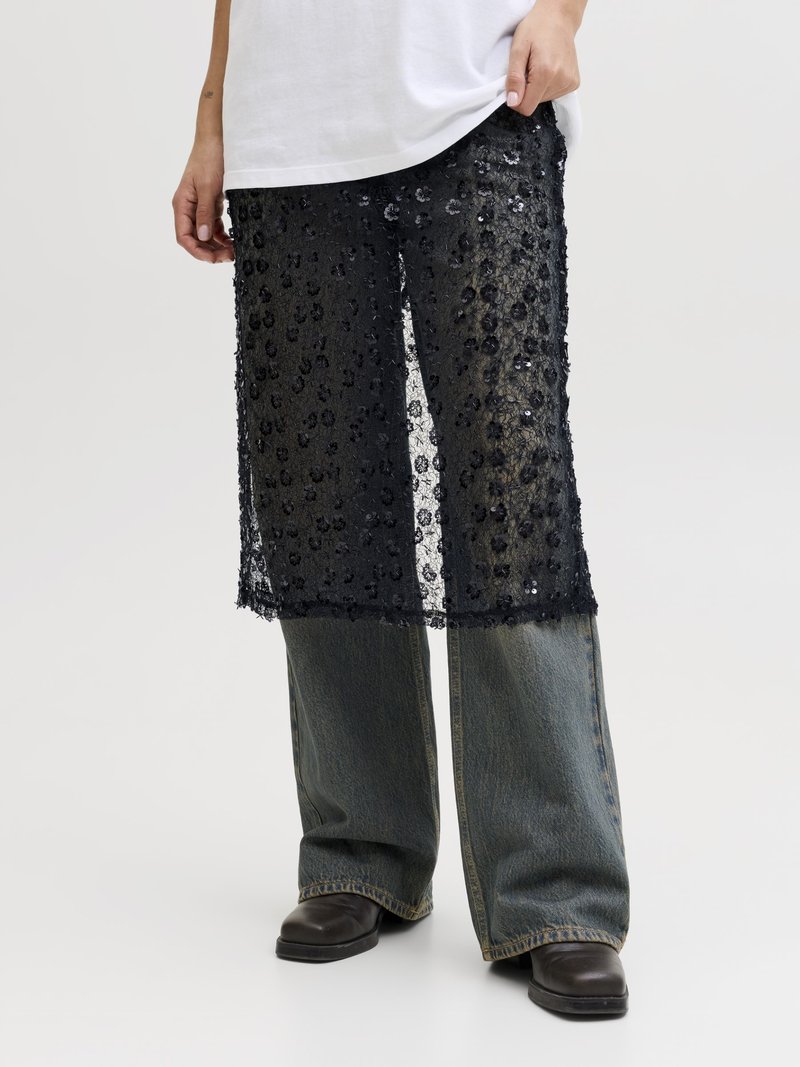 Person trägt einen schwarzen, mit Pailletten besetzten, transparenten Rock, der über weit geschnittene Jeans und schwarze Schuhe gelegt ist, und hält mit einer Hand einen weißen Hemdsaum fest.
