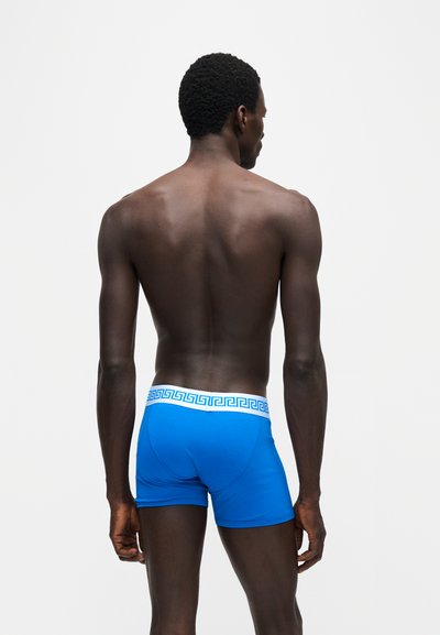 Homme à la peau foncée et aux cheveux courts bouclés, debout de dos, portant un boxer bleu vif avec une ceinture blanche ornée d’un motif géométrique bleu.