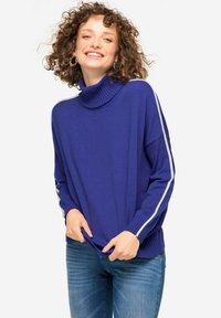 Maglione a collo alto viola con texture a coste, caratterizzato da strisce bianche lungo le maniche, abbinato a jeans azzurri chiari.