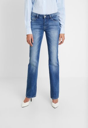 Person trägt blaue Jeans mit geradem Bein, hellblau gestreiftes Hemd und weiße spitze Absatzschuhe und steht vor einem schlichten Hintergrund.