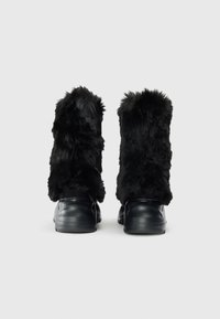 Botas de pelo negro con un upper texturizado y esponjoso y suelas de goma negra lisa. El diseño tiene una altura corta con un destacado detalle de pelo.