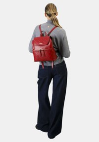 Zaino in pelle rossa con motivo a rilievo, tasca frontale e spallacci regolabili. Il modello indossa un maglione grigio e jeans a gamba larga.