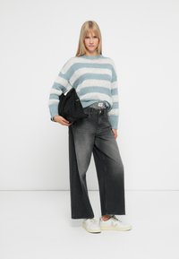 Kvinne iført en blå og hvit striped genser, vide, falmede svarte jeans, hvite joggesko og holder en svart veske mot en hvit bakgrunn.