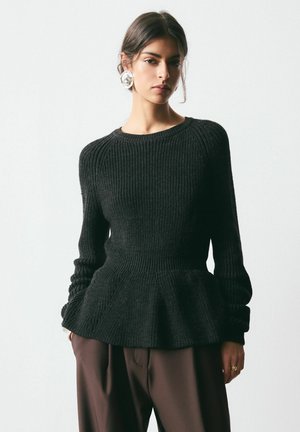 Žena v čiernom rebrovanom pletenom svetri s volánom, hnedej nohavice, veľkých strieborných kruhových náušniciach a zlatom prsteni stojaca pred jednoduchým svetlým pozadím.