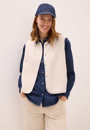 Beige fleece-vestillä, jossa on teksturoitu pinta, napitus, yhdistettynä siniseen farkkupaitaan ja vastaavaan siniseen lippikseen. Neutraali tausta.