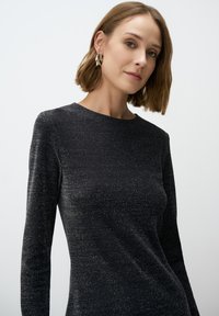 Abito lungo nero con una sottile texture scintillante, scollatura rotonda e silhouette aderente. La modella indossa grandi orecchini a cerchio.