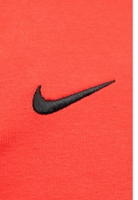 Červená tkanina Nike obsahuje čierne vyšívané logo Swoosh, ktoré zdôrazňuje jej hladkú textúru a žiarivú farbu.
