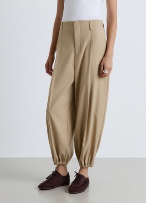 Persona che indossa pantaloni beige a vita alta a sigaretta con polsini elastici e scarpe marrone scuro con lacci, in piedi su un pavimento chiaro contro una parete bianca.