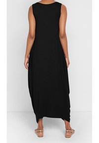 Czarna, bezrękawkowa sukienka maxi o luźnym kroju, wykonana z miękkiego materiału; posiada okrągły dekolt i drapowane boki, zestawiona z płaskimi sandałami.