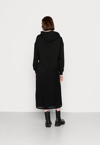 Hoodie noir avec une capuche à cordon, associé à une jupe plissée noire. La tenue est complétée par des bottines noires à la cheville.