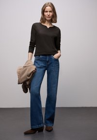 Ciemna bluzka z długim rękawem i dekoltem w serek, zestawiona z jasnoniebieskimi dzwonowatymi dżinsami. Modelka trzyma beżową kurtkę, nosi brązowe, spiczaste botki za kostkę.