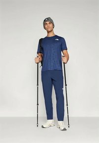 Jauns vīrietis jūras zila sporta apģērbā un pelēkā cepurē tur pārgājienu stabi, mugurā viņam ir melna mugursoma un baltas sporta kedas, un viņš ir pozējis pret vienkāršu fona.