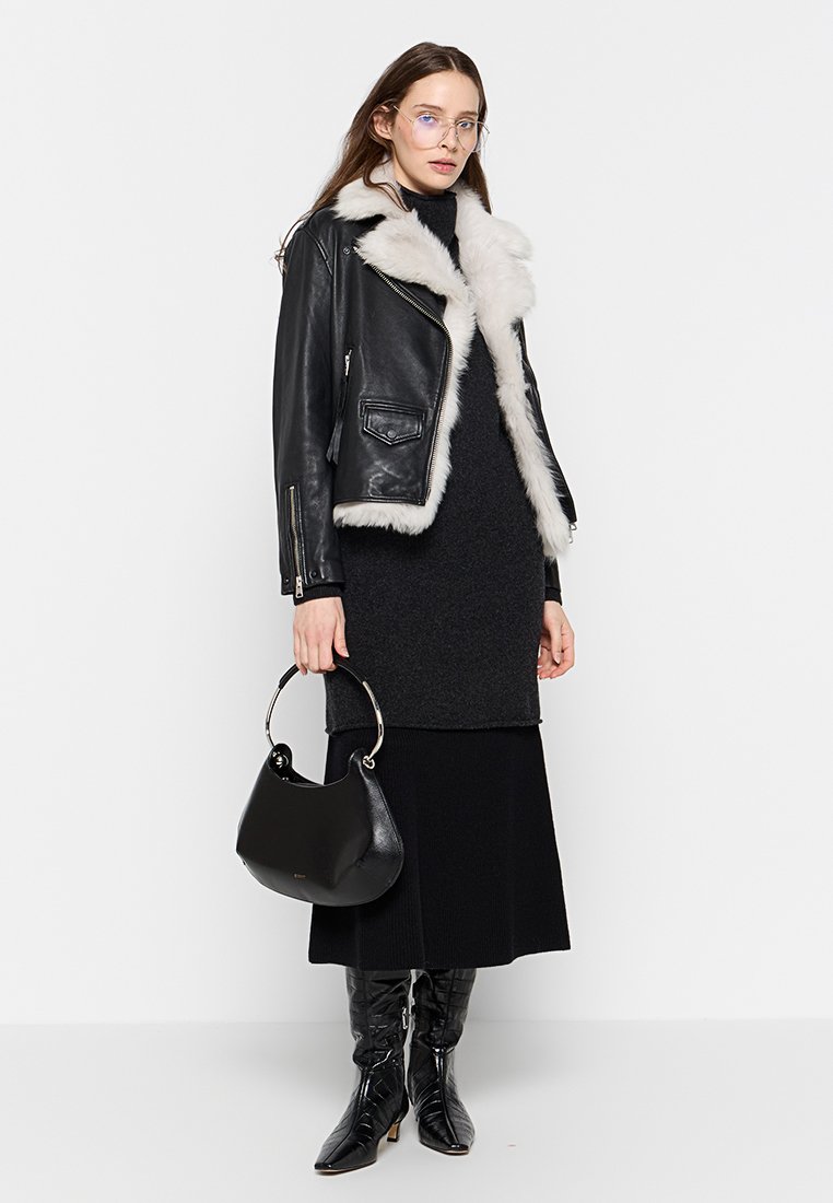 Schwarze Leder-Bikerjacke mit weißem Pelzkragen, kombiniert mit einem dunklen Strickkleid, schwarzen kniehohen Stiefeln und einer strukturierten schwarzen Handtasche.