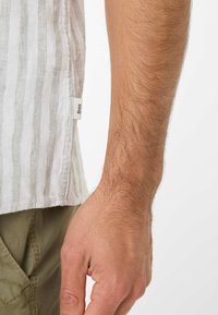 Camicia a maniche corte, bianca a righe, realizzata in tessuto leggero; etichette visibili; abbinata a pantaloni verde oliva. Primo piano di braccio e mano.