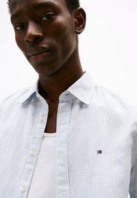 Camisa de botones a rayas en azul claro con cuello, con botones blancos y un pequeño logo bordado en el pecho. Camiseta interior blanca visible.