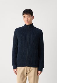 Suéter de punto acanalado azul marino oscuro con cremallera, cuello alto y puños ajustados, hecho de un material texturizado. Combina con pantalones beige claros.