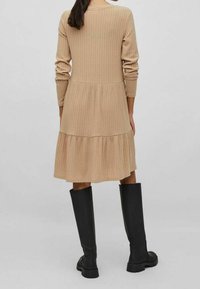 Robe beige côtelée longueur genoux avec manches longues et ourlet à volants. Associée à des bottes noires montant jusqu'aux genoux avec une semelle épaisse.