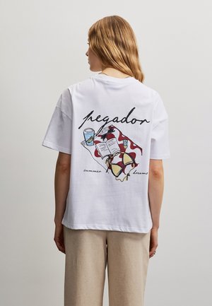Világosbarna hajú nő, aki fehér pólót visel, amelyen nyári témájú grafika látható: bikini, ital, könyv és törölköző a "pegador" és „summer dreams” felirattal.