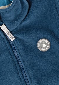 Marineblaue Fleecejacke mit hohem Kragen, vollem Reißverschluss und einem runden grauen Logoakzent. Sie verfügt über eine weiche Textur und einen kontrastierenden Reißverschluss.
