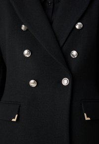 Blazer negro de corte doble con tejido texturizado. Presenta botones decorativos plateados y acentos dorados en los bolsillos.