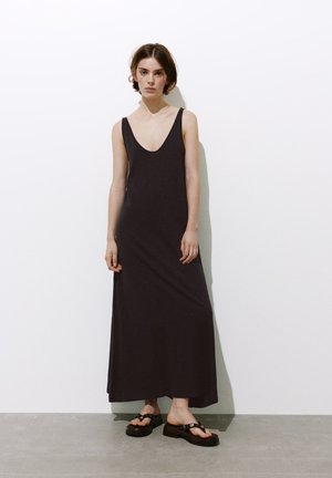 Jeune femme debout contre un mur blanc, portant une robe maxi noire sans manches et des sandales à plateforme noires avec des détails cloutés.