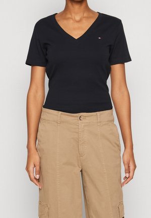 Vrouw draagt een zwart T-shirt met V-hals en korte mouwen met een klein logo en beige cargobroek met voorzakken en een knoopsluiting.