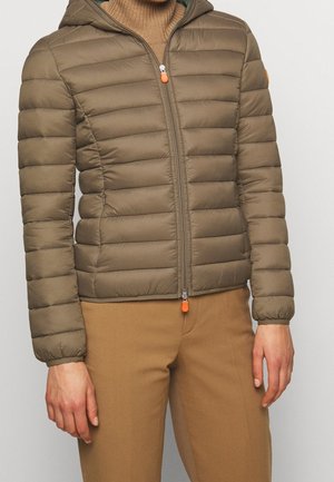 Personne portant une veste matelassée à capuche marron fermée par une fermeture éclair, avec un pantalon marron assorti et un pull à col roulé marron clair en dessous.