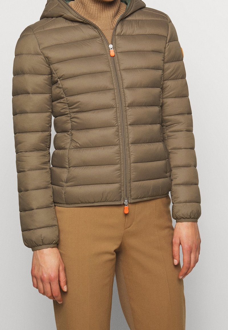 Personne portant une veste matelassée à capuche marron fermée par une fermeture éclair, avec un pantalon marron assorti et un pull à col roulé marron clair en dessous.