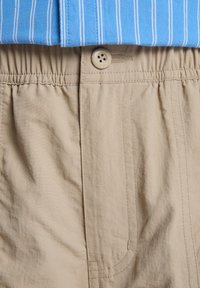 Close-up van beige broek met elastische tailleband, knoopsluiting en gulp, gecombineerd met een blauw-wit gestreept overhemd dat is ingestopt.
