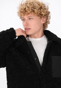 Giacca in pile nera e fluffy con superficie testurizzata, chiusura con bottoni e tasca a toppa. Indossata sopra una camicia bianca, che mette in mostra un design elegante.