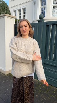 Smėlio spalvos megztinis su pintine rašta ir plačiomis rankovėmis, derinamas su blizgios faktūros leopardžio rašto sijonu. Lauko aplinka su žalia tvora.